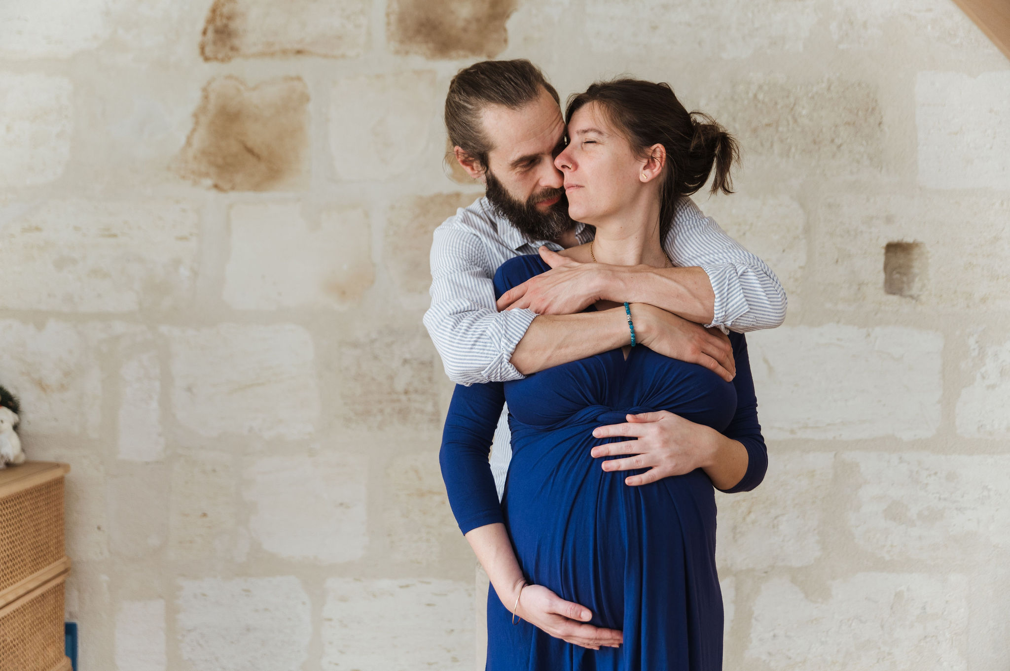 Futur papa enlaçant tendrement sa compagne enceinte devant un mur en pierre