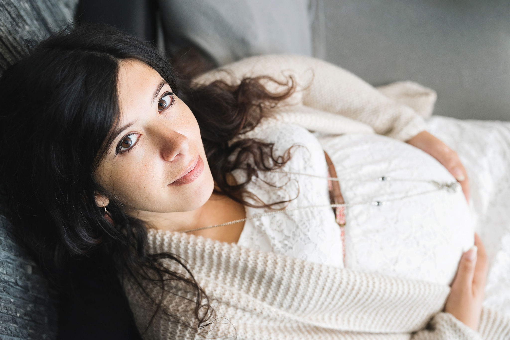 Future maman allongée chez elle, regard vers l'objectif, séance grossesse à domicile