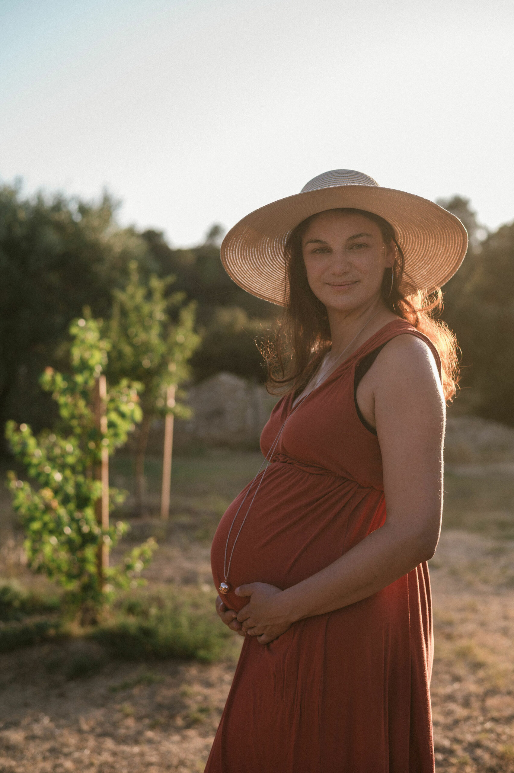 Future maman en robe rouge et chapeau, en lumière de fin de journée, séance grossesse près d'Orléans